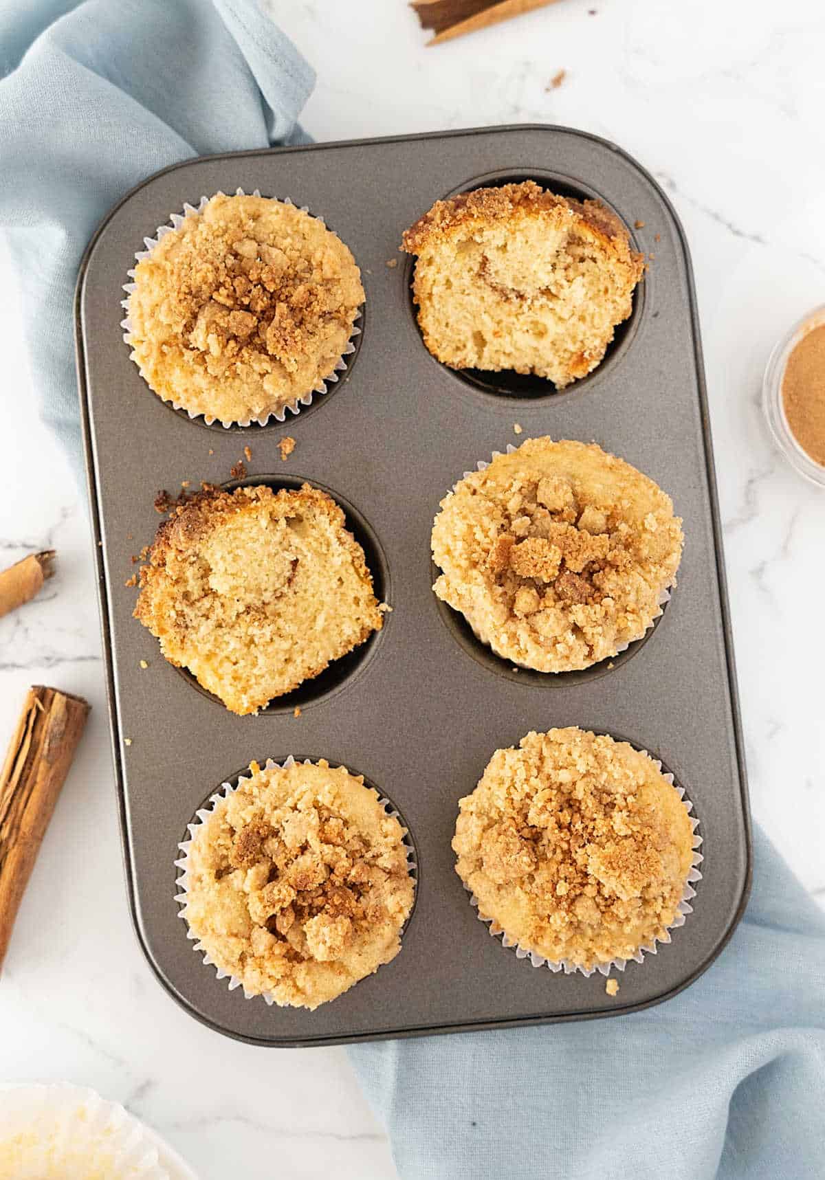 Metal pan with whole and halved cinnamon crumb muffins. Light blue cloth, white marbled surface. 