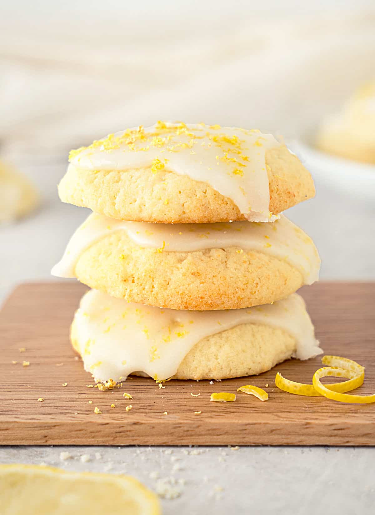 Wooden board with stack of three glazed lemon cookies. Grey surface, beige background.