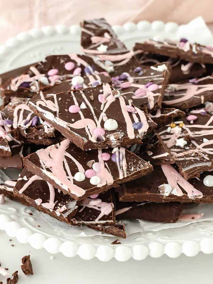 A white plate with pile of dark chocolate and pink bark pieces. Light pink background.