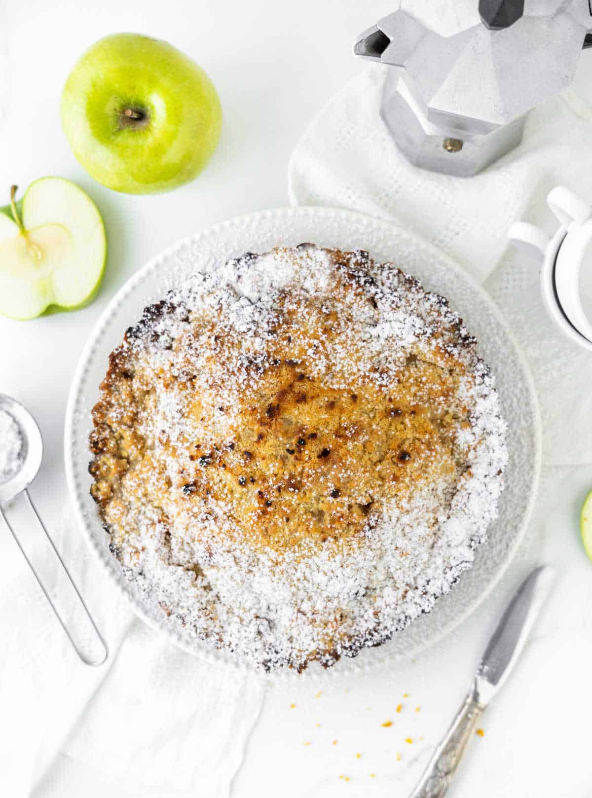 Whole apple crumb pie on a white surface. Green apples, cutlery, a metal coffee pot. Top view.