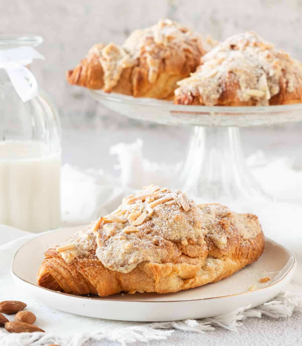 Glass pie plate with almond croissants and white plate with single one. White and gray background. Milk bottle. 