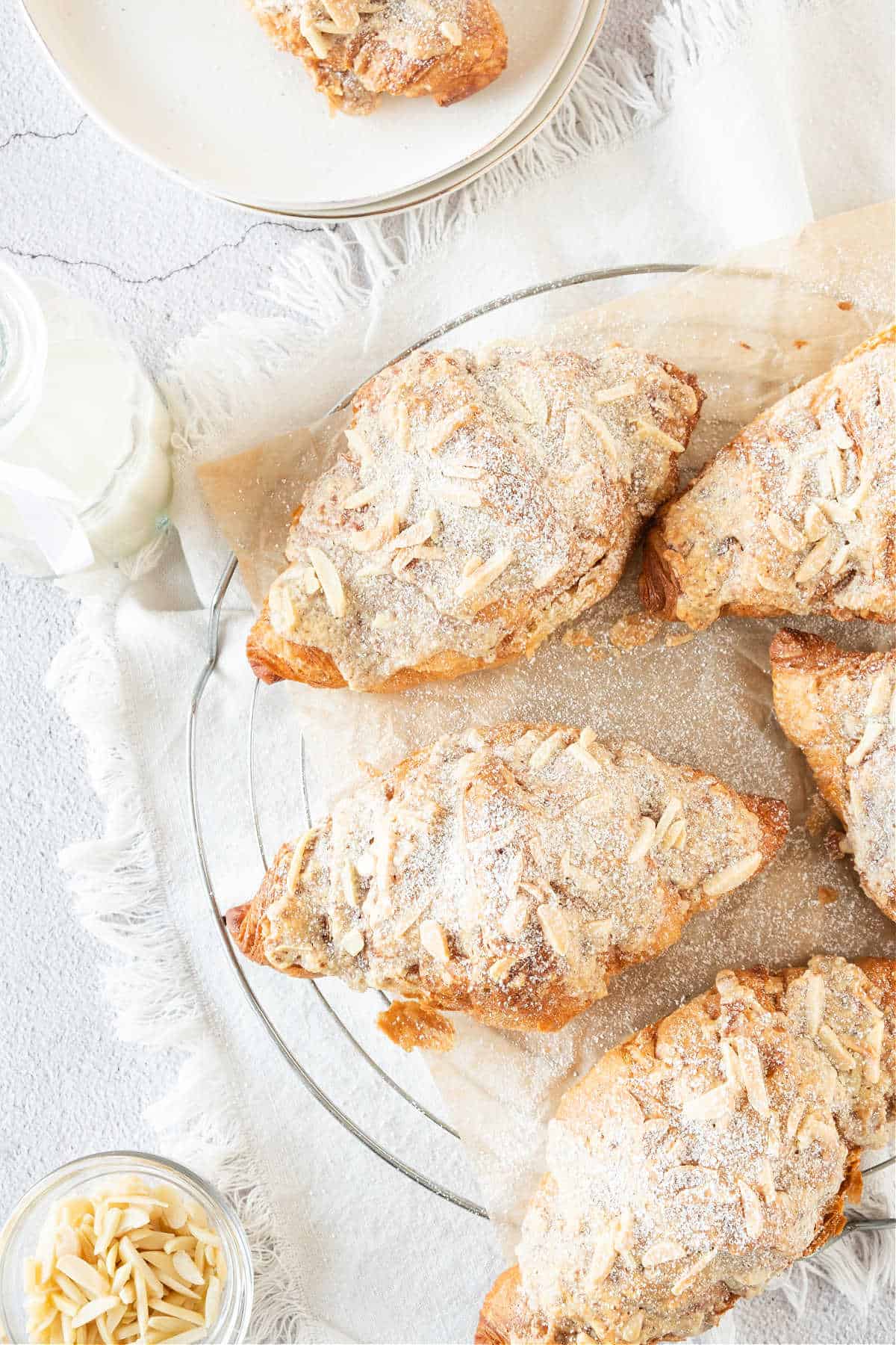 Top view of several almond croissants on a white plate with a white cloth. Light gray surface. 