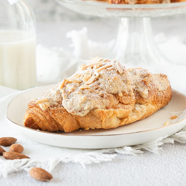 Single almond croissant on a white plate. Light gray surface, white cloth, glass bottle in the background.