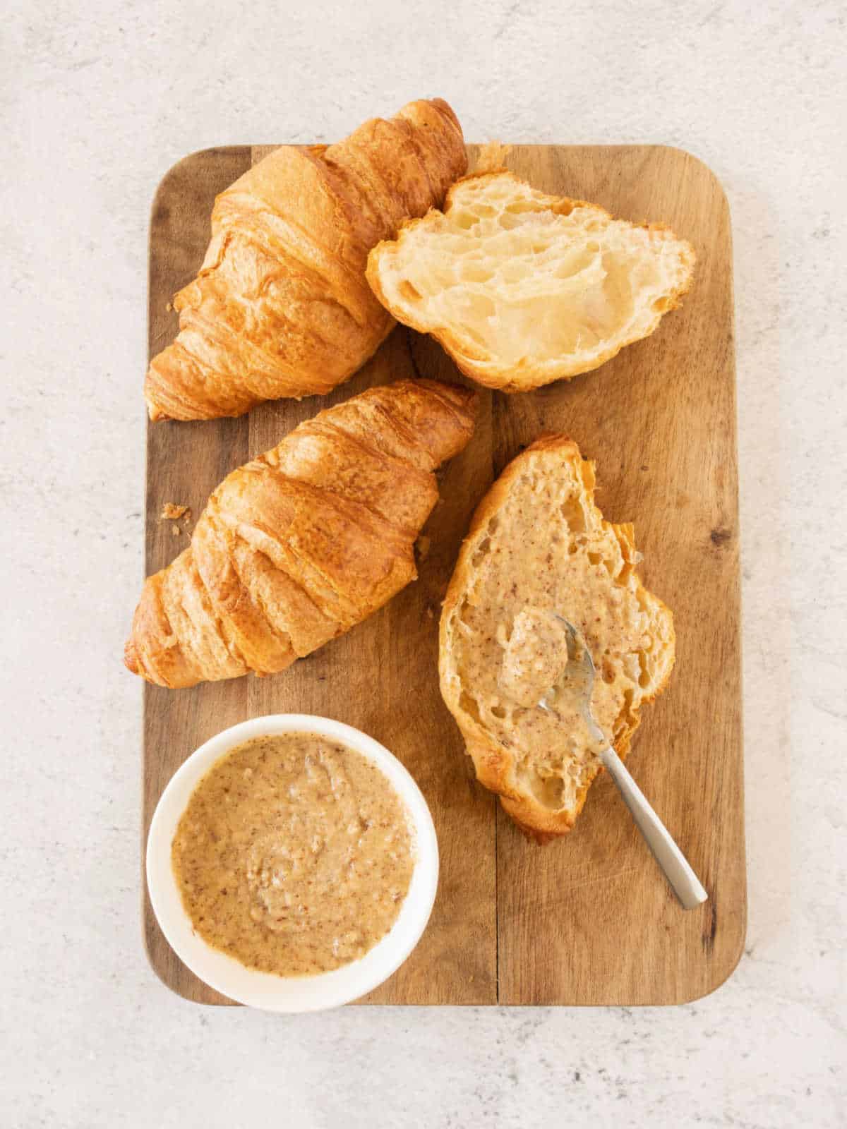 Spreading almond cream on a half croissant on a wooden board with whole croissants. Light gray surface.