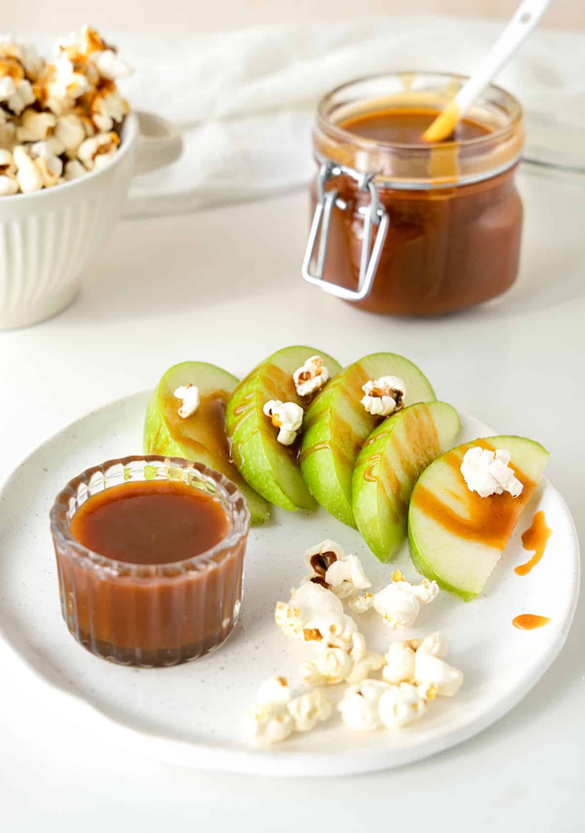 White plate with green apple slices, popcorn and caramel. White background with caramel jar.