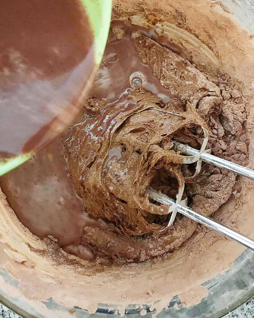 Adding a liquid chocolate mixture to a chocolate batter in bowl with beaters.