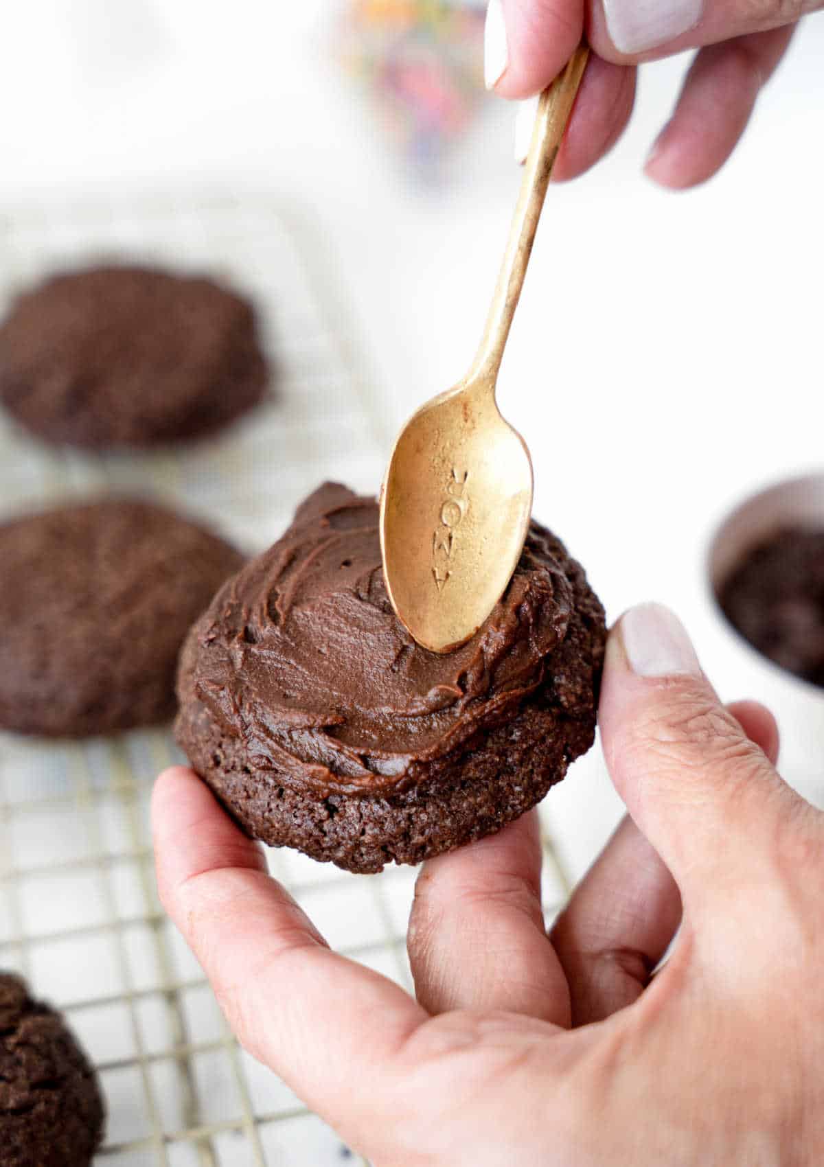 Frosting a chocolate cookie with ganache and a golden spoon. More cookies below. White background.