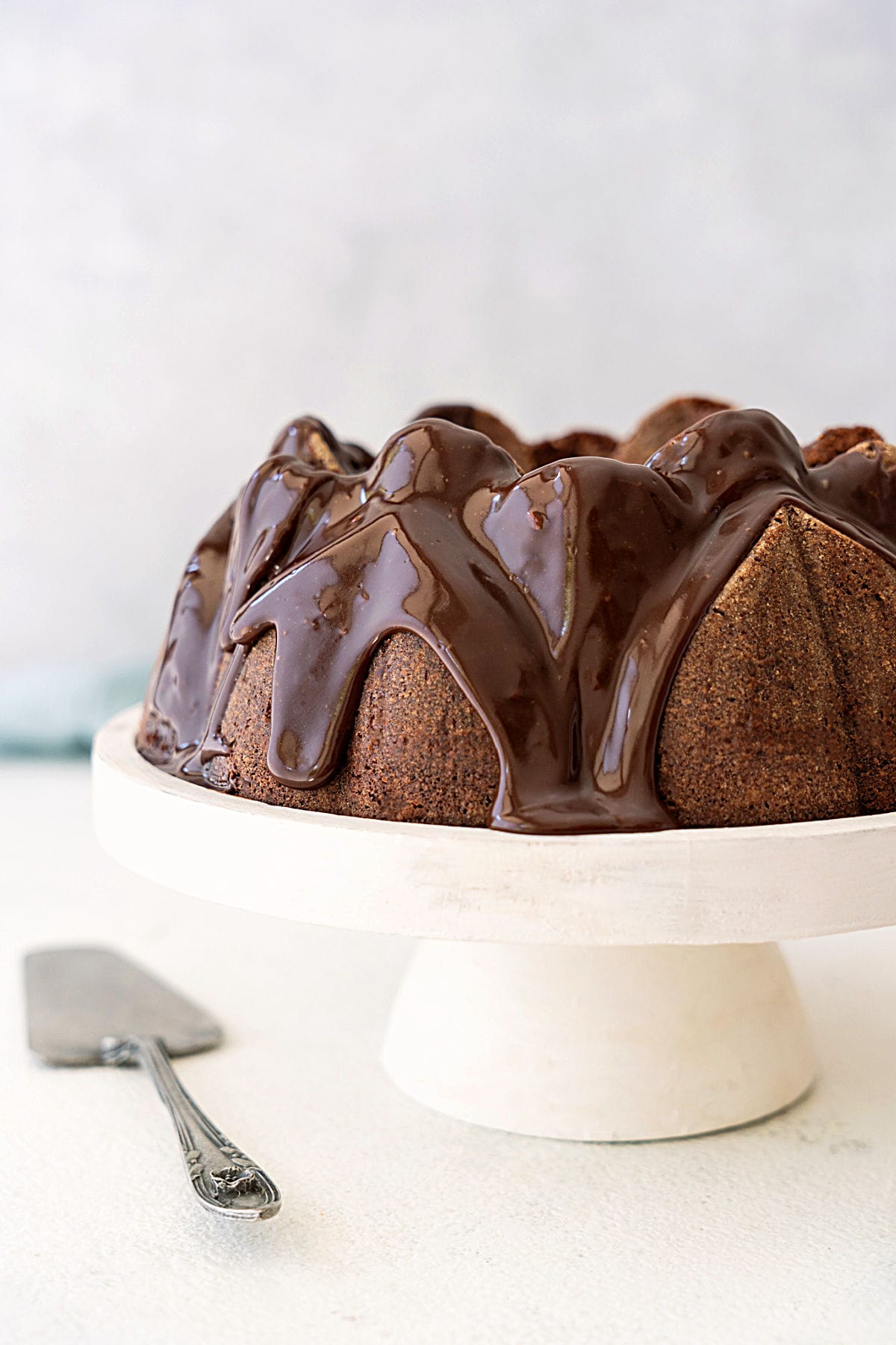 Partial view of glazed chocolate bundt cake on a white cake stand. White surface, light gray background.