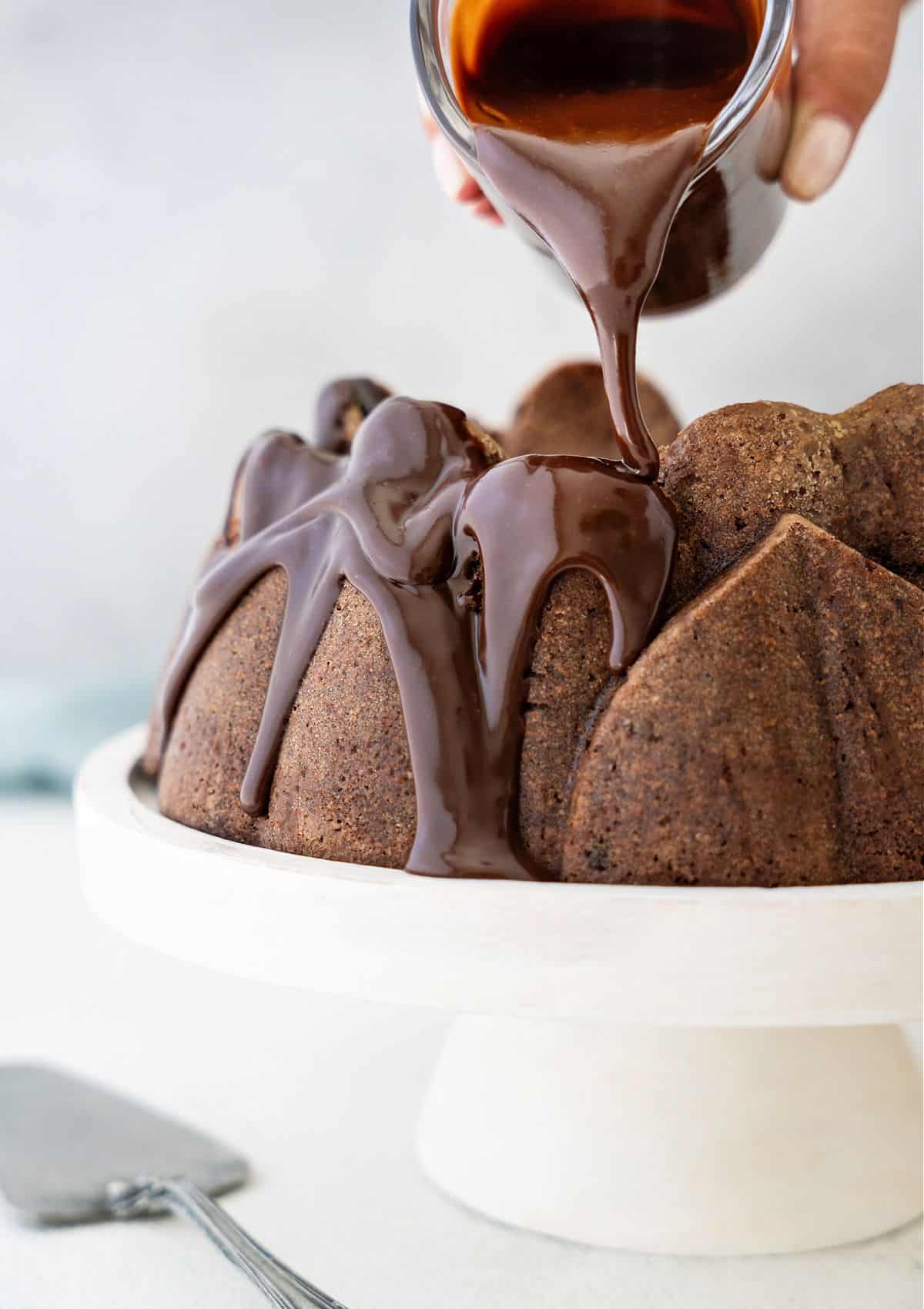 Glazing a chocolate bundt cake on a white cake stand. Light grey background.