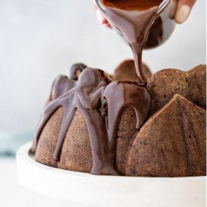 White plate with chocolate bundt cake being glazed. Light gray background.