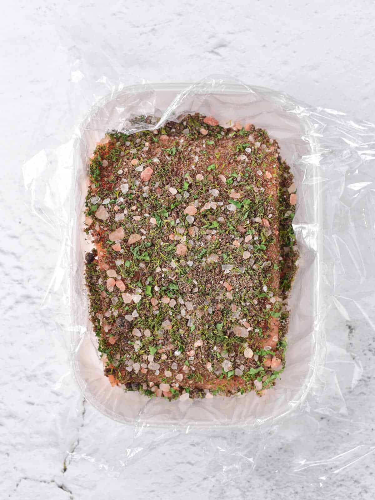 Plastic lined container with salmon covered in salt and herbs. White marble surface.