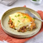 Serving of shepherd's pie on a green plate. Silver fork, clay-colored cloth, light gray surface.