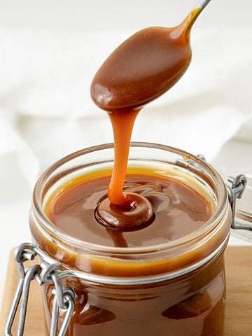 Spooning caramel sauce from a mason jar on a wooden board. White background.