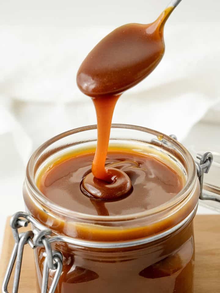 Spooning caramel sauce from a mason jar on a wooden board. White background.
