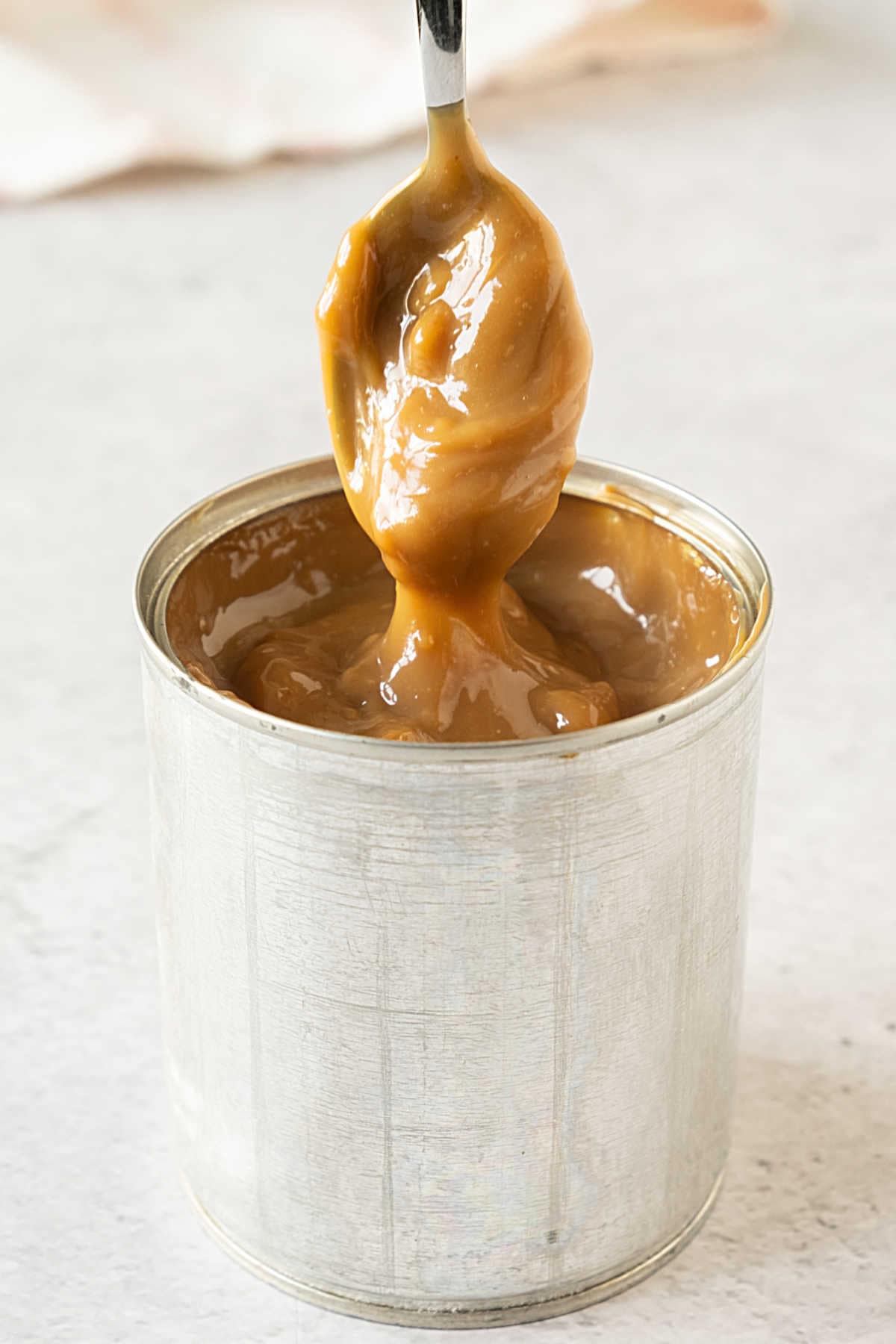 Lifting spoon with dulce de leche from a metal can on a light gray surface. 