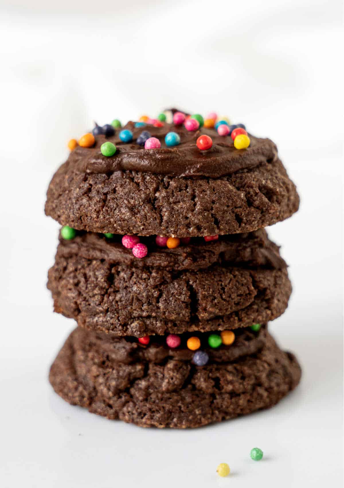 Stack of three frosted chocolate cookies with round colorful sprinkles. White marble surface. 