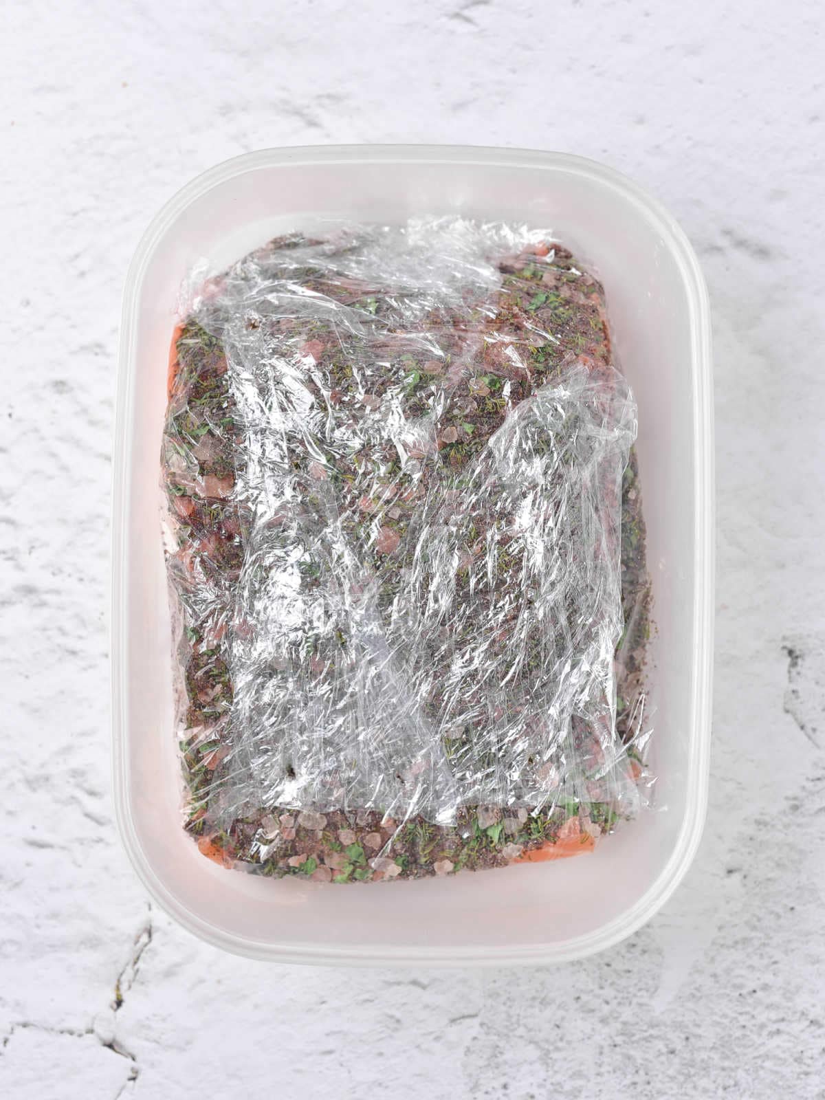 Light gray surface with white baking dish containing plastic-wrapped salt-crusted salmon piece.