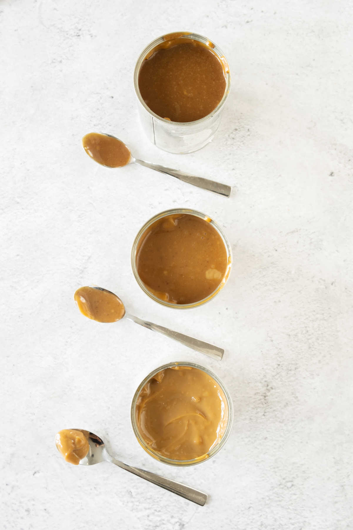 Top view of three cans and spoon with dulce de leche on a light gray surface.