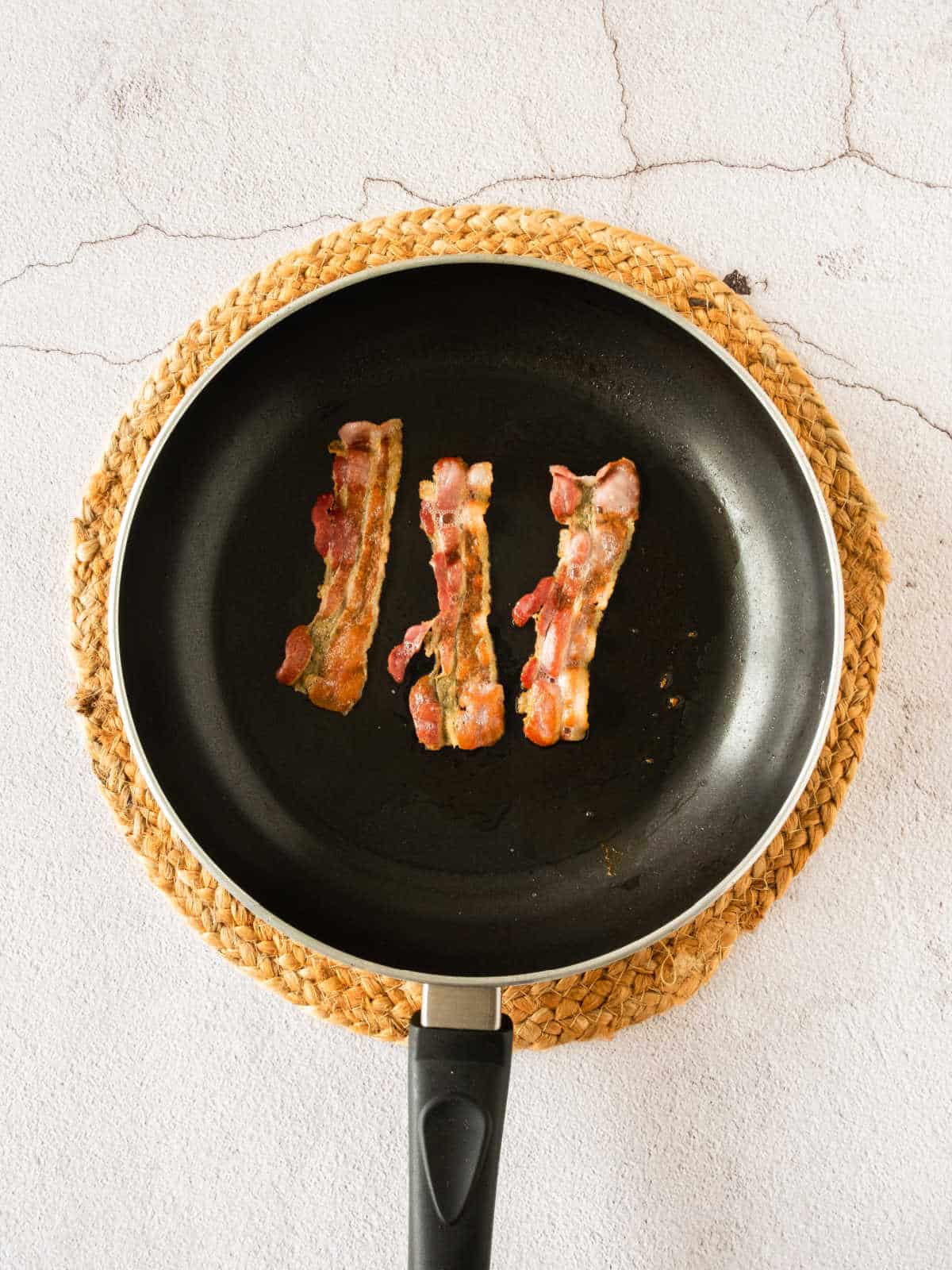 Crispy bacon slices in a black skillet on a beige placemat. Light gray surface.