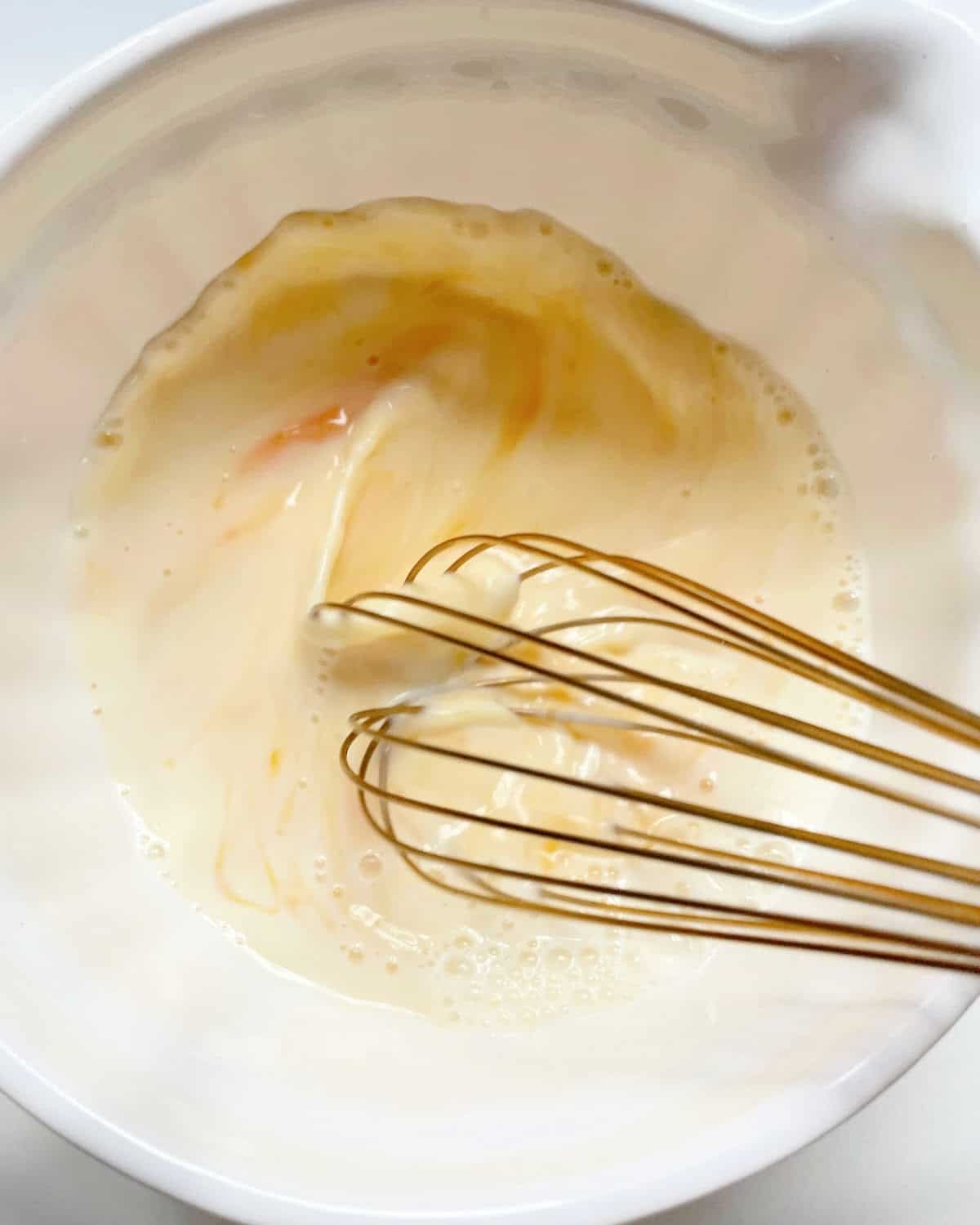 Whisking eggs and milk in a white bowl with a gold-colored whisk.