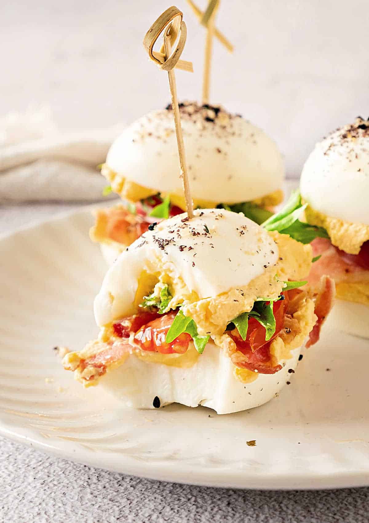 Bitten skewered BLT deviled egg sandwiches on a white plate. White surface and background.