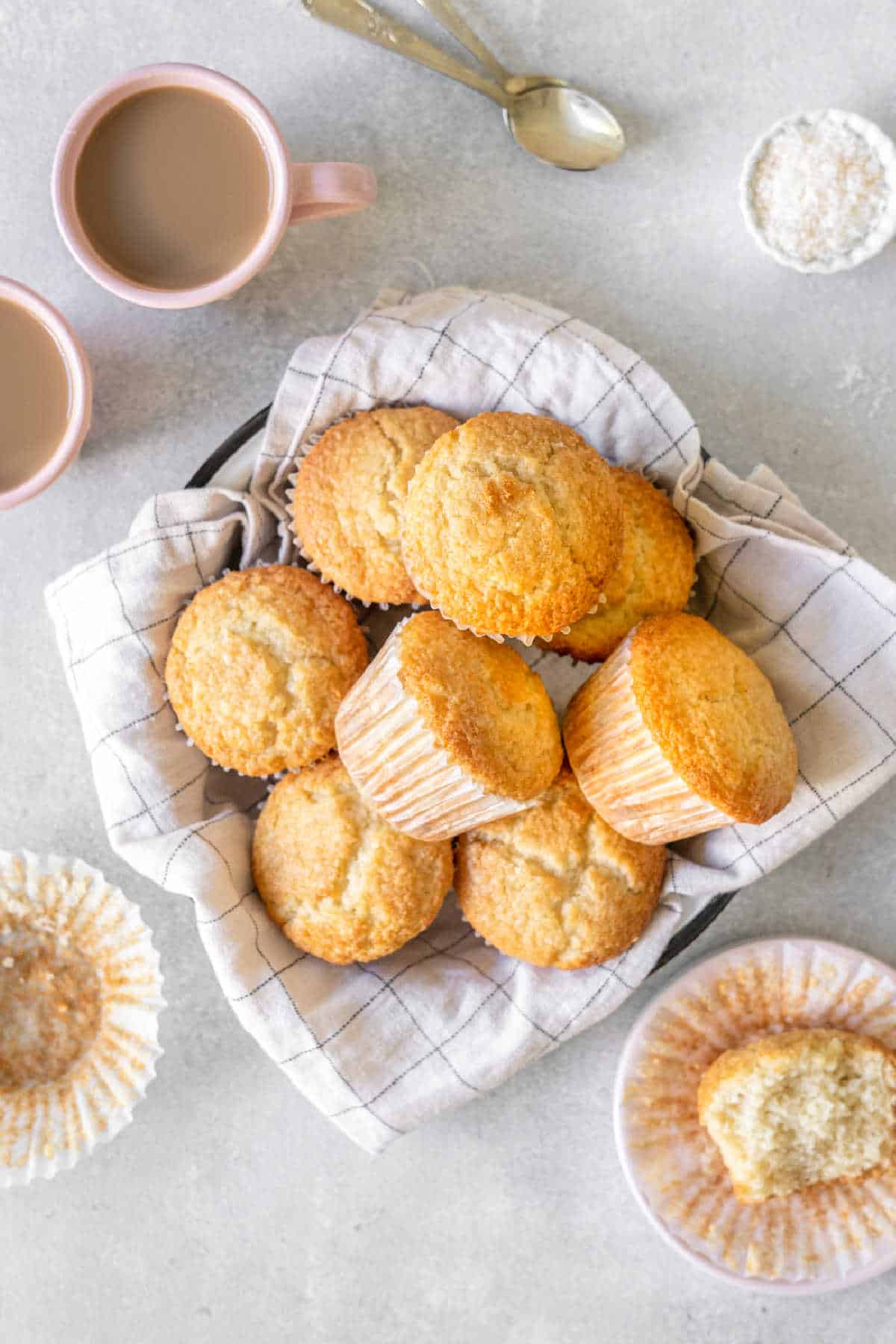 Pile of coconut muffins on a white checkered cloth. Light gray background. Coffee mugs. Top view.