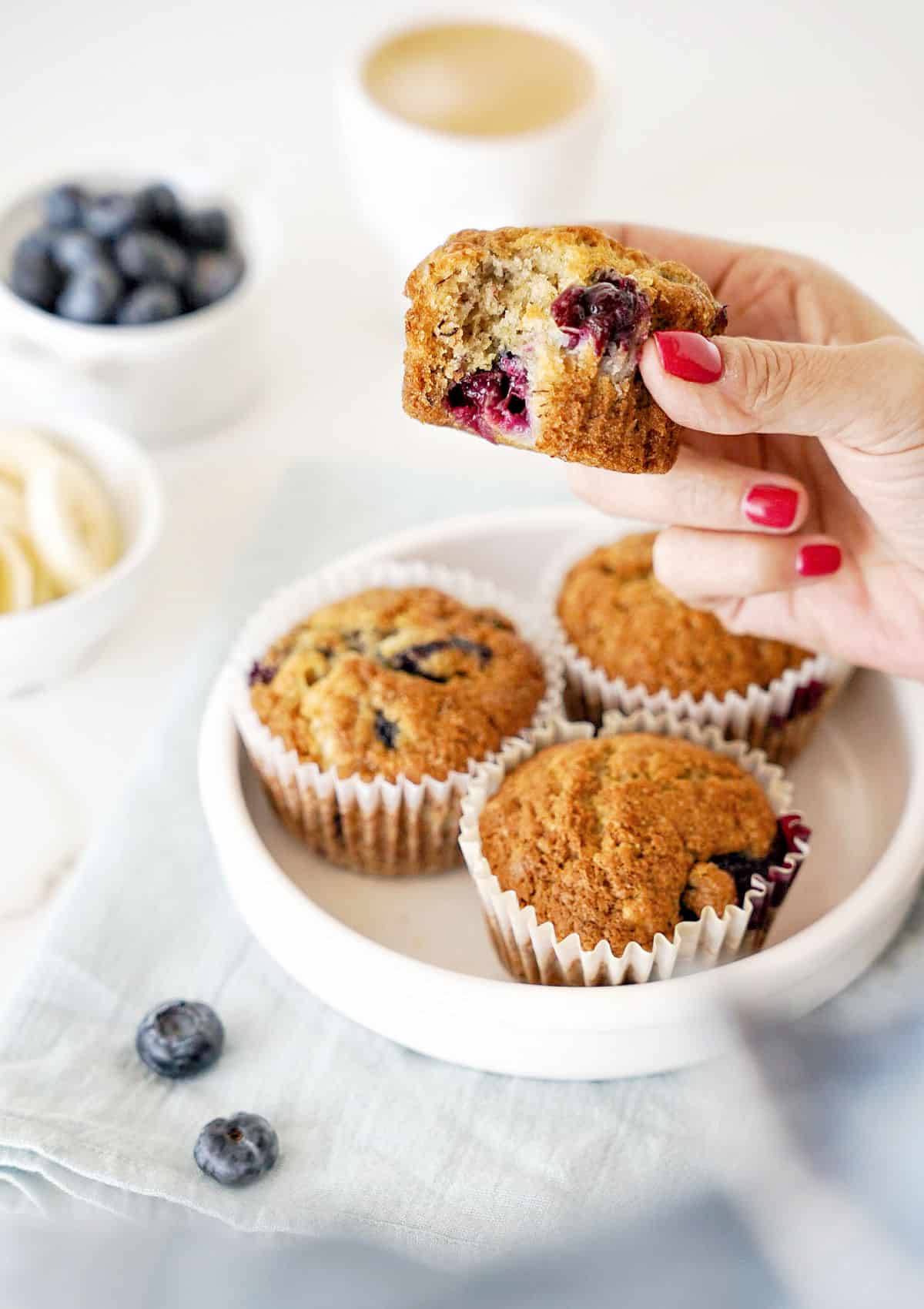 Holding a bitten blueberry banana muffin over white plate with wole ones. White surface with fruit in bowls.