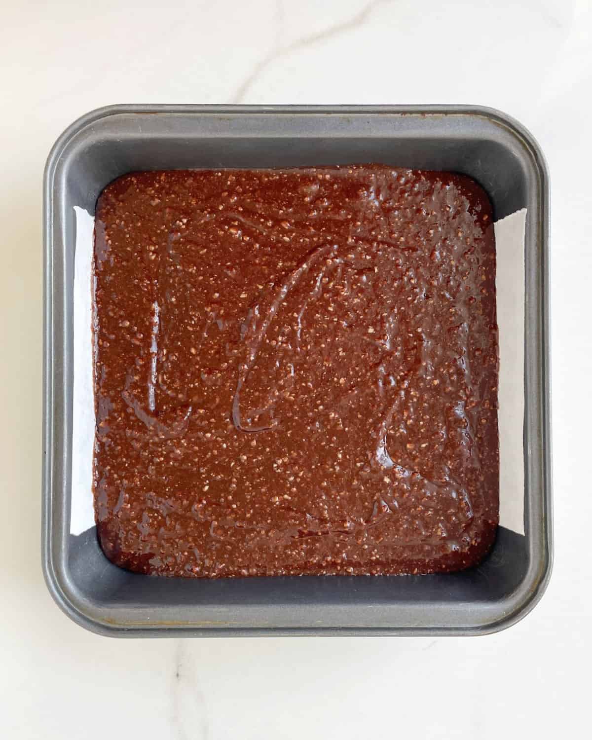 Brownie batter in a square metal pan on a white marble surface.