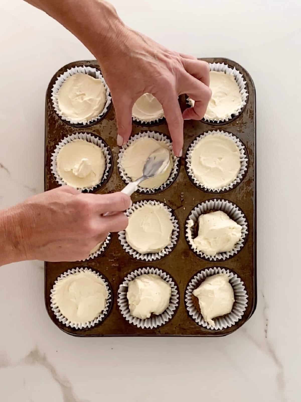 Filling paper liners in a muffin pan with no bake cheesecake. White marbled surface.