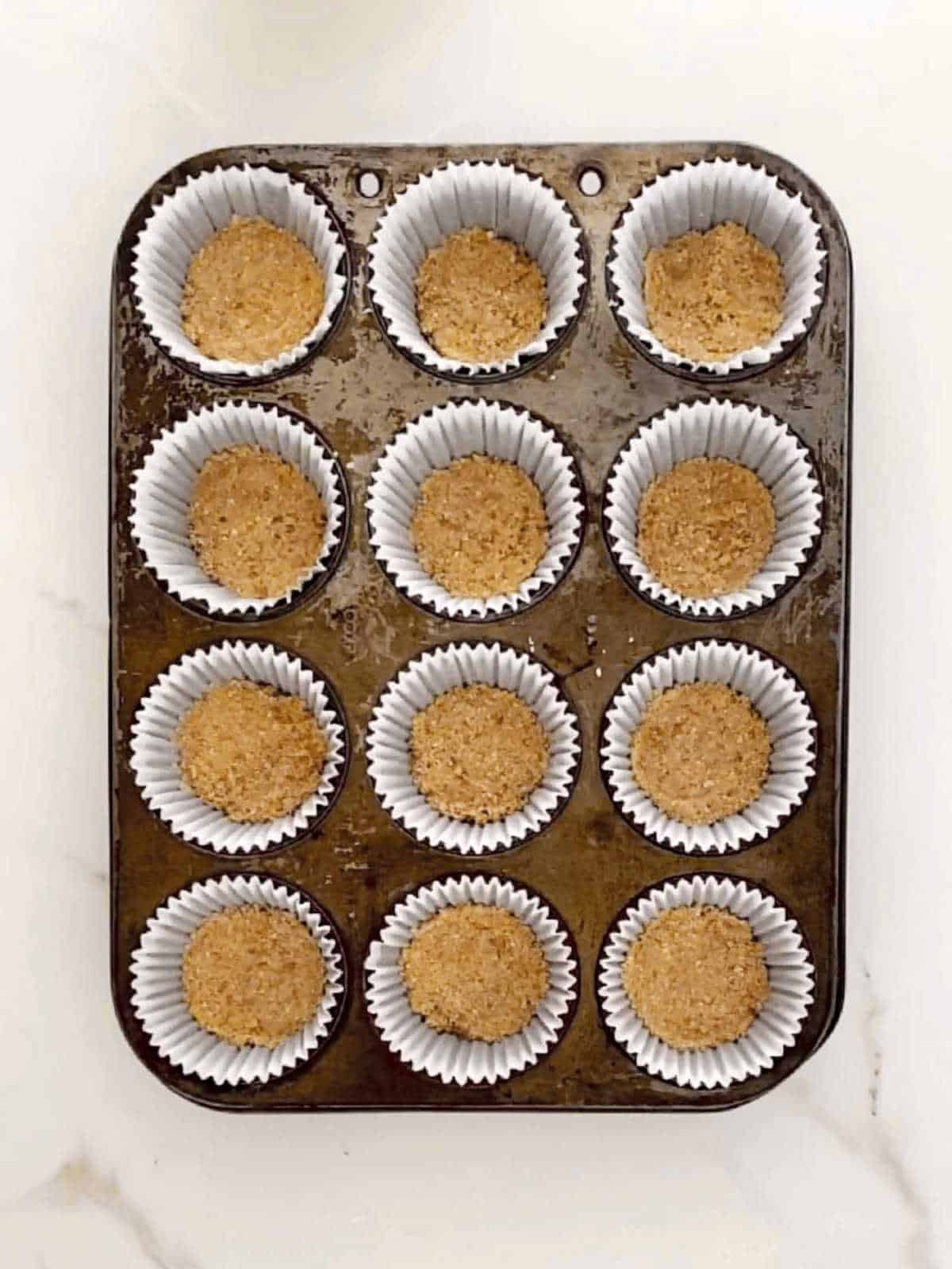 Metal muffin pan with paper liners and pressed crumb crust. White marbled surface.