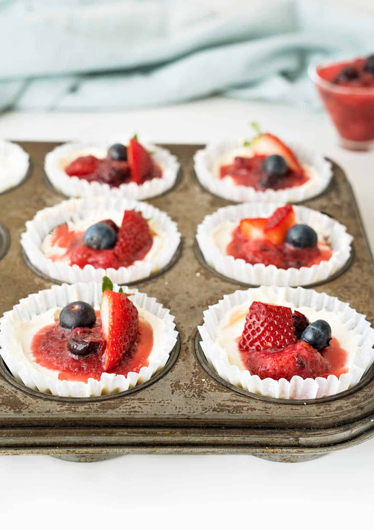 Two rows of mini berry cheesecakes in paper liner in a muffin tin. Light blue background.