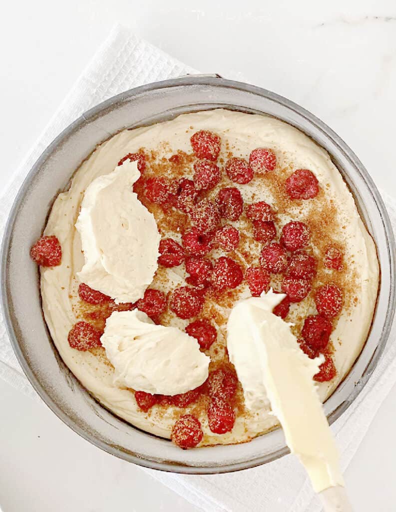 Mounding cake batter with a spatula over raspberries in a cake pan. White surface.