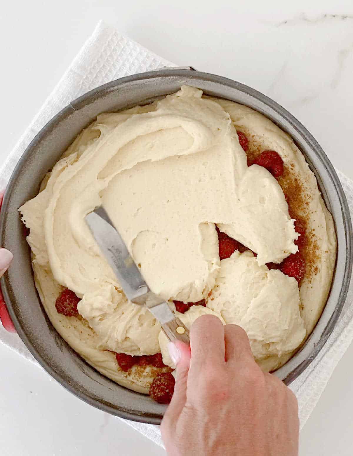 Spreading cake batter over raspberries in a round cake pan on a white surface.