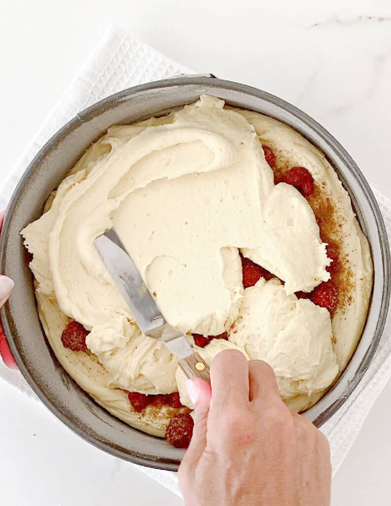 Spreading cake batter over raspberries in a round cake pan on a white surface.