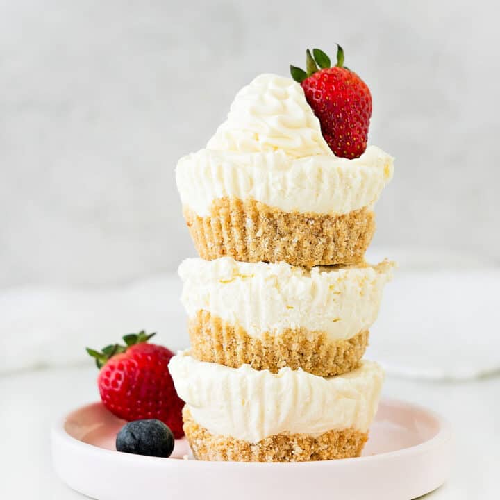 Light gray background with stack of three mini cheesecakes on a pink plate. Berries. White surface.