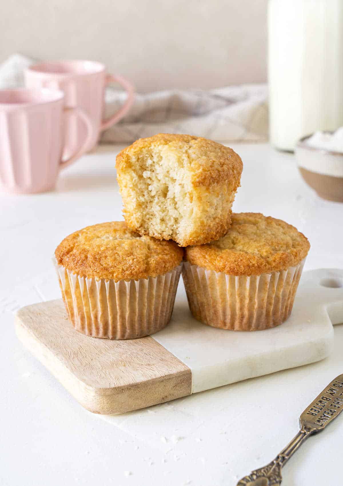 Bitten coconut muffin on top of two whole ones on a board. White surface. Grey, pink and white background. 
