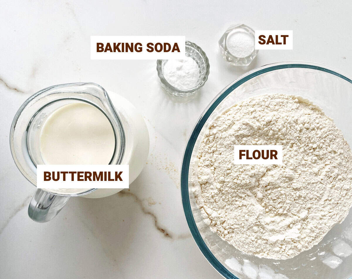 White marble surface with bowls containing ingredients for soda bread: buttermilk, flour, salt and baking soda.
