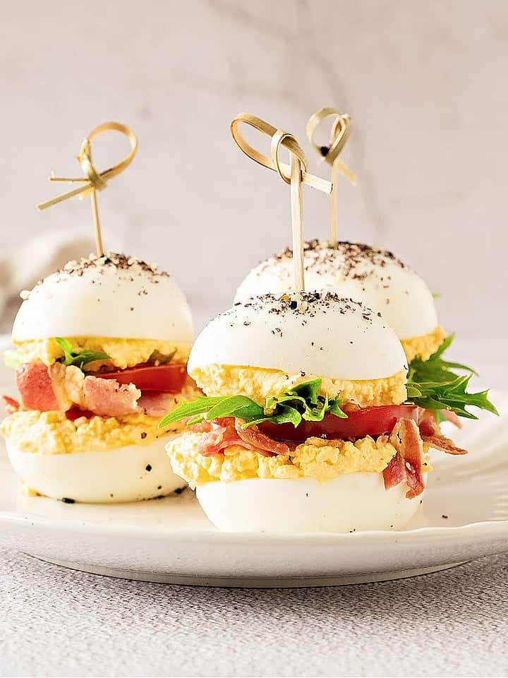 Three BLT deviled egg sandwiches on a white plate. Whitish grey surface and background.