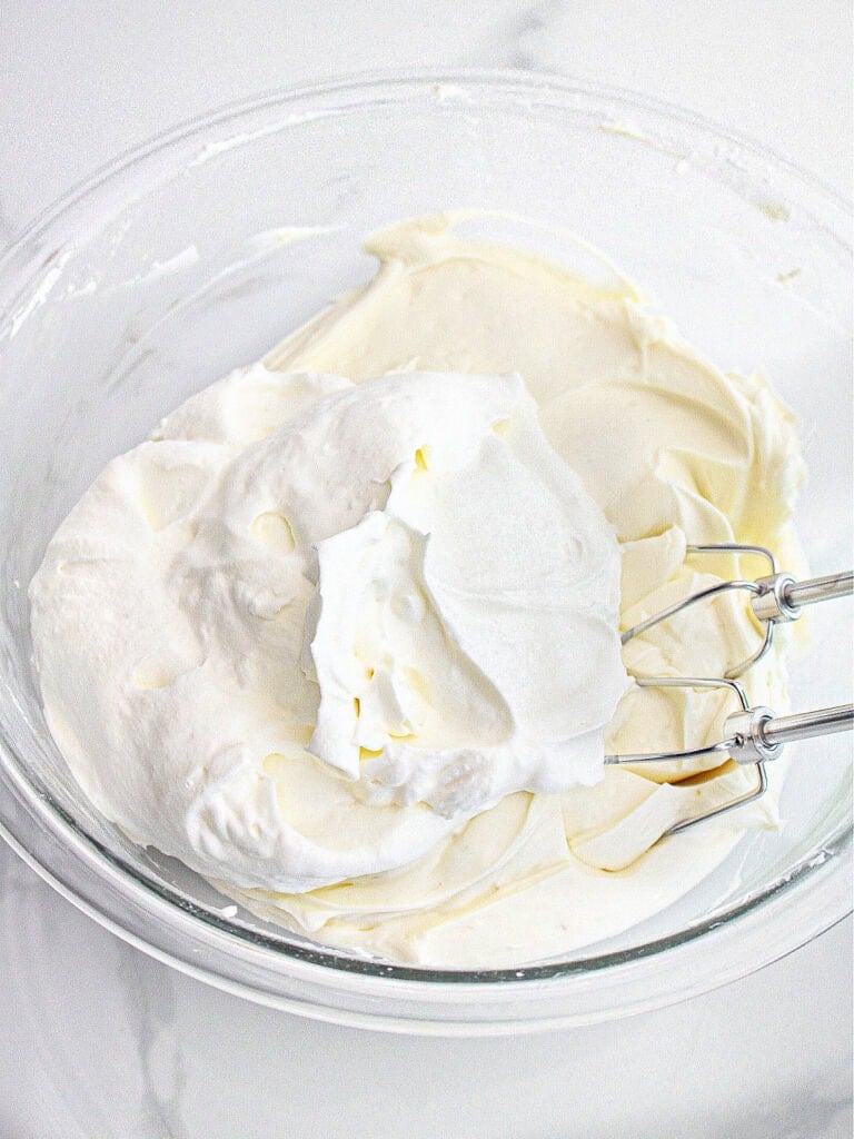 Cream cheese and whipped cream in a glass bowl with metal beaters. White marble background.