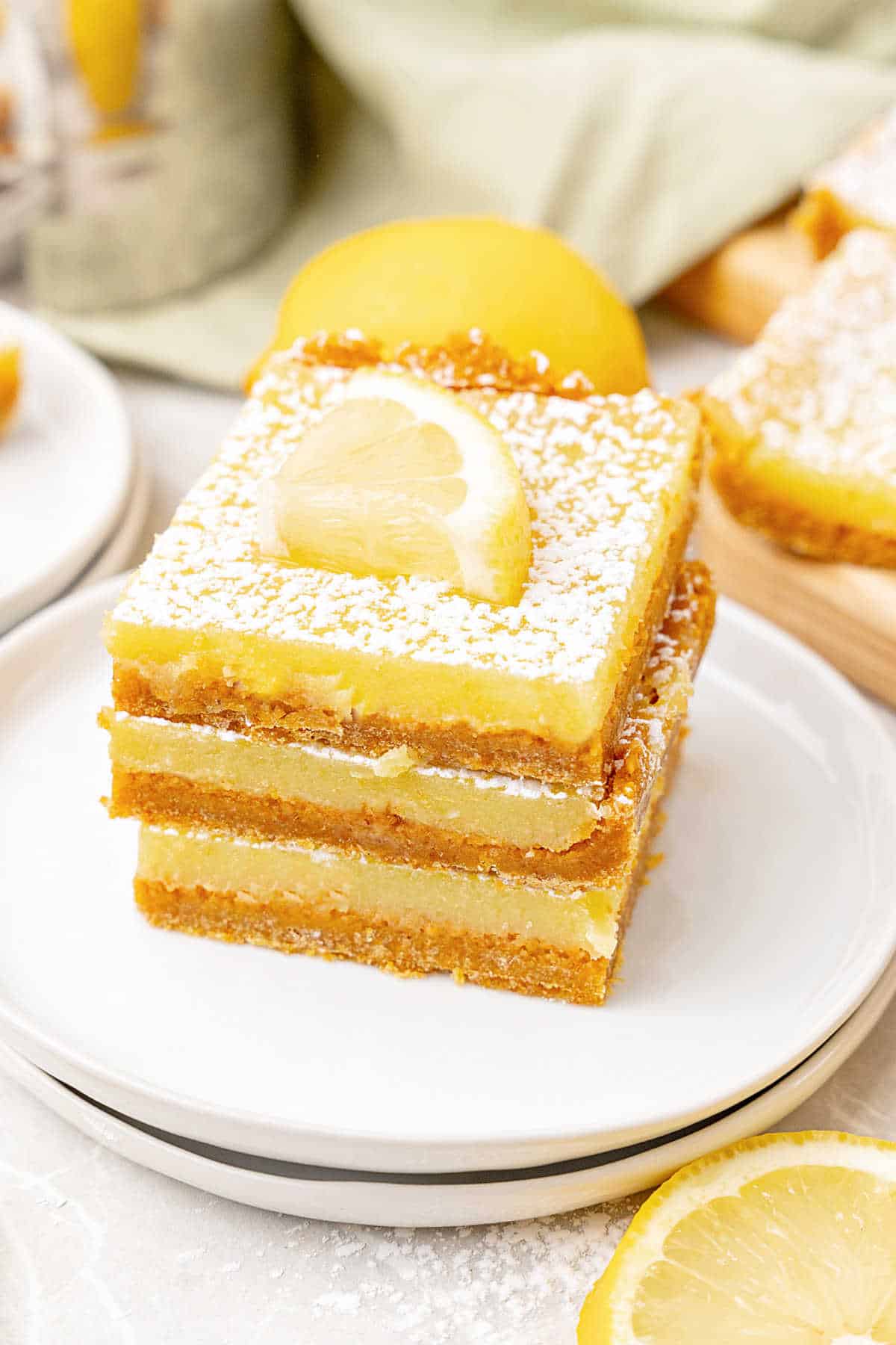 Three lemon bars on a stack of white plates. White surface with lemon slices, metal cup, green cloth.