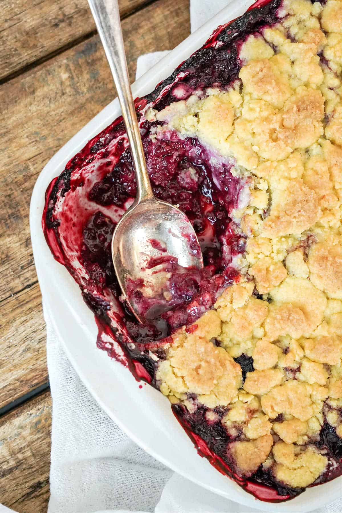 Silver spoon in a white dish with berry dump cake. Wooden table, white linen.