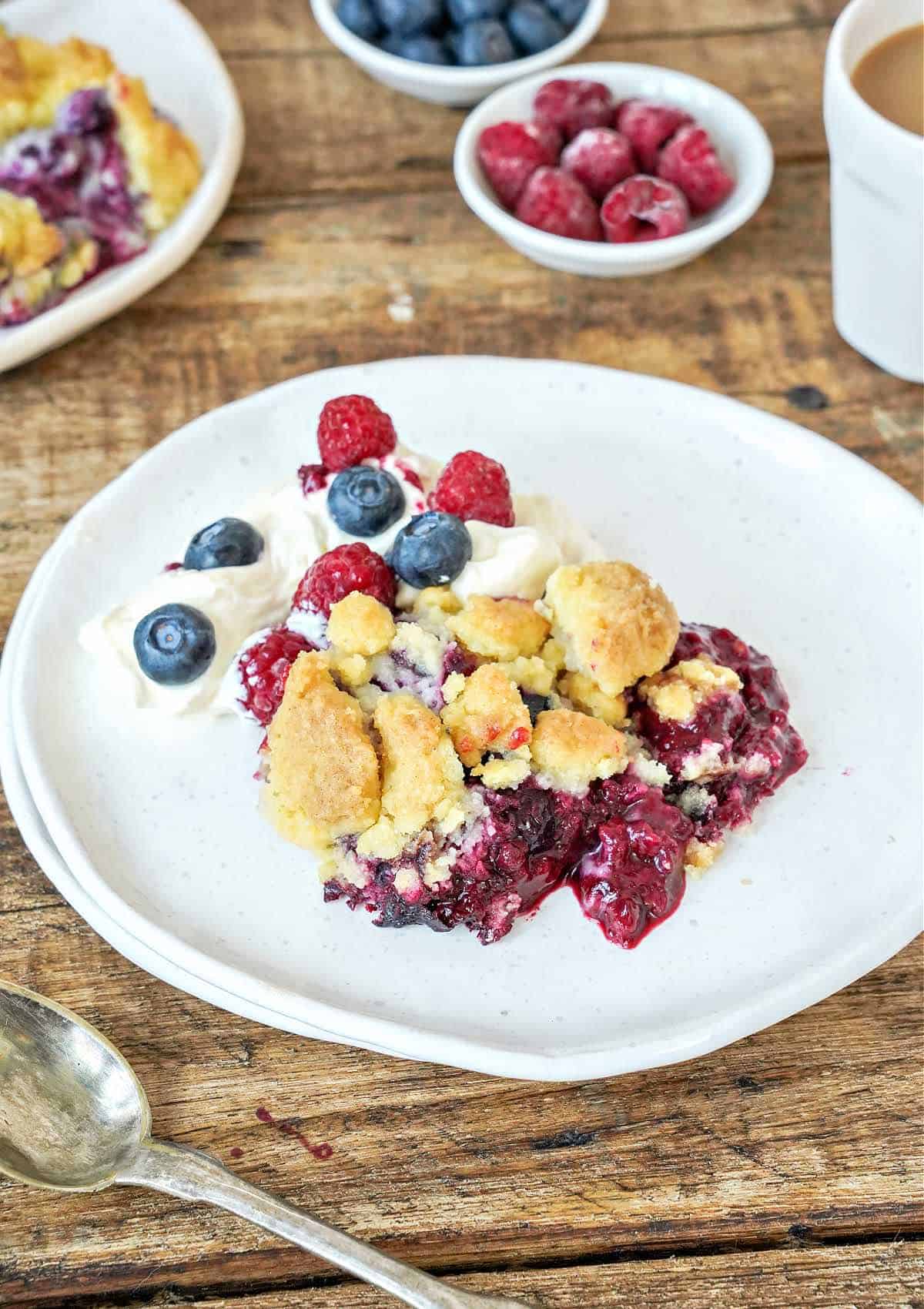 White plate with serving of mixed berry dump cake. Wooden table. Fruit in bowls.