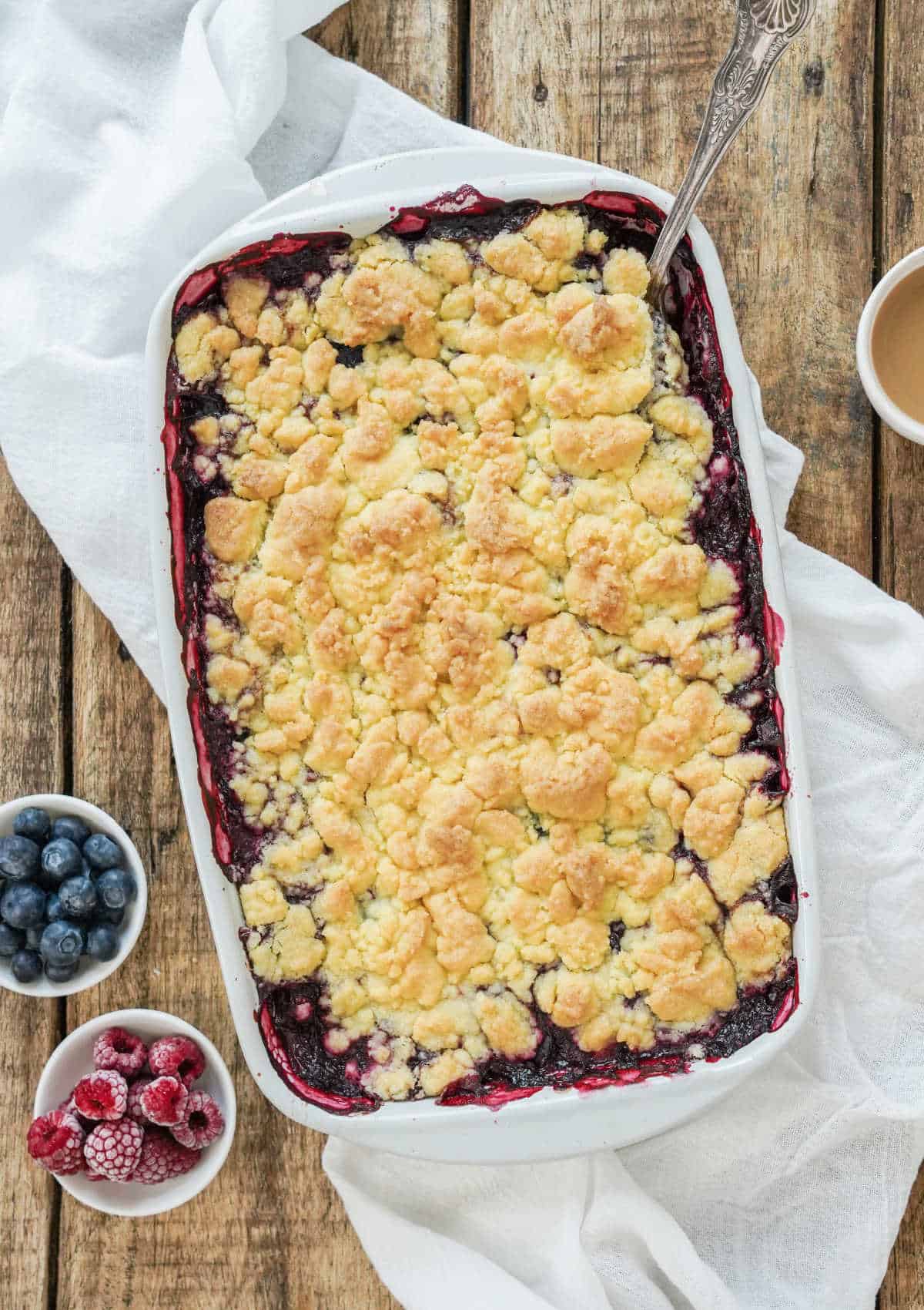 Top view of rectangular white baking dish with berry dump cake. White cloth, wooden table, bowls of berries.