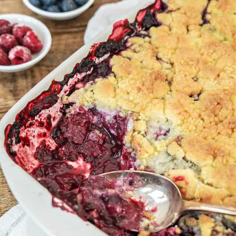 Close up of baking dish with berry dump cobbler. Silver spoon, wooden surface, bowls with berries.
