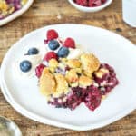 Mixed berry dump cake with cream on a white plate. Wooden surface.