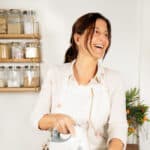 Person with apron laughing in a kitchen with electric mixer in hand.