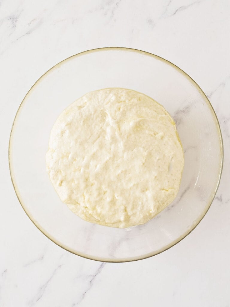 Glass bowl with bread dough before rising. White marbled background.