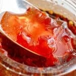 White and brown text overlay on jar and spoon of red chili jam.