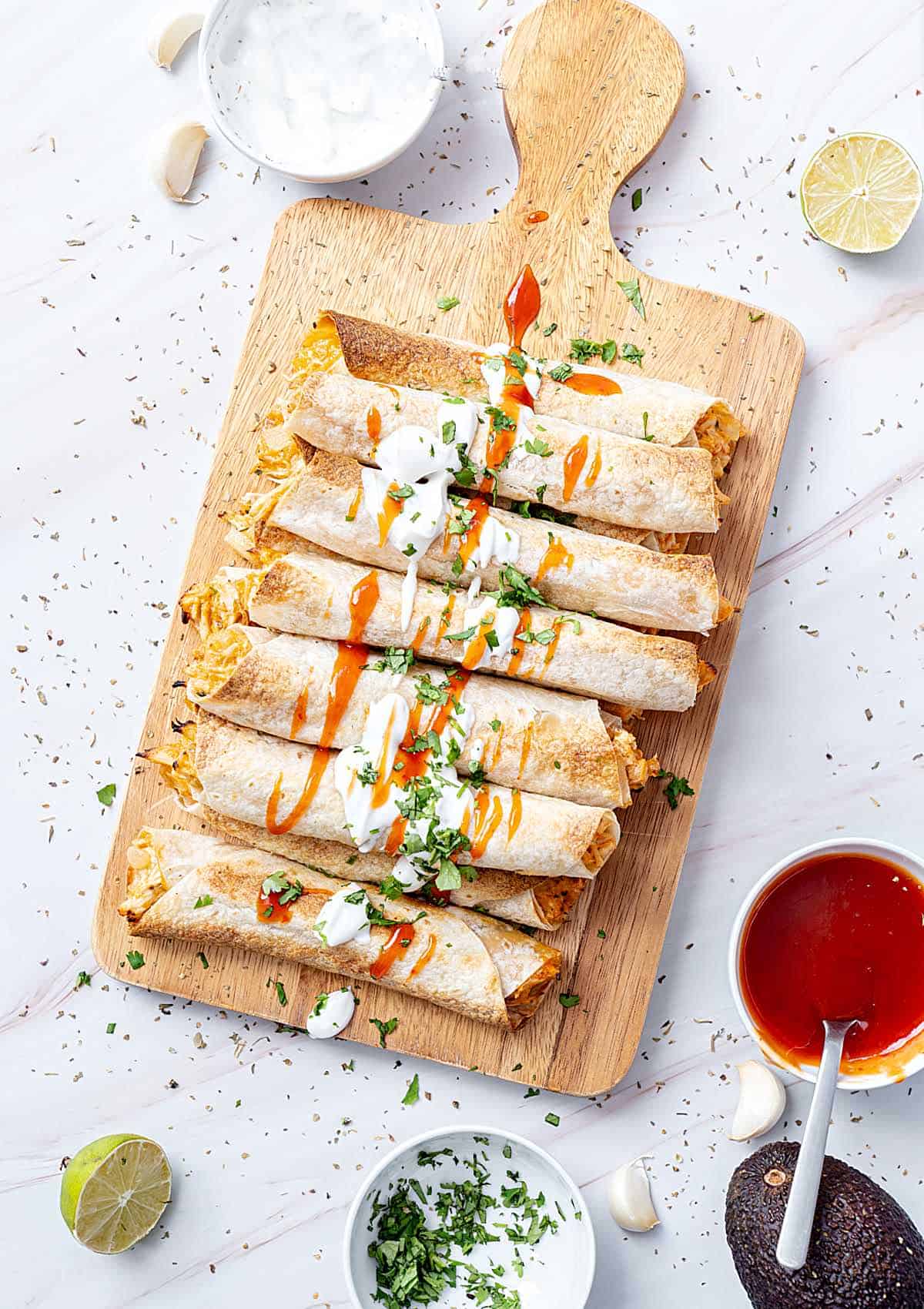 White marbled surface with wooden board and chicken taquitos. Bowls with sauce and cream. Top view. 