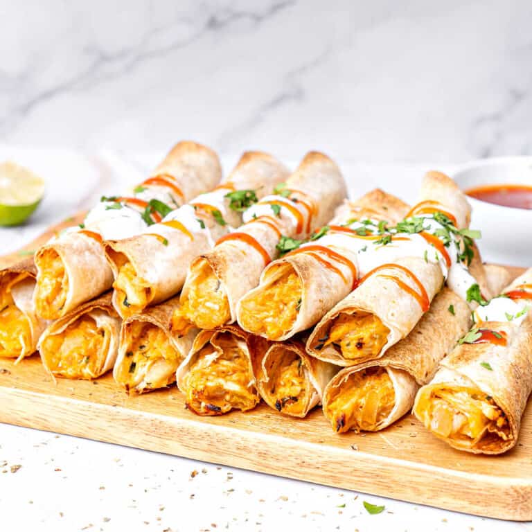 Two rows of piled baked chicken taquitos on a wooden board. White marbled background.